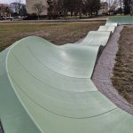 20260319_115507 Zielony tor pumptrack na tle budynków i drzew w parku. Tło wypełniają drzewa i niskie zabudowania. Pogoda jest słoneczna z lekkim zachmurzeniem.