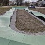 20260319_115513 Zielony, falisty tor pumptrack otoczony jest żwirem. W tle stoją drzewa i budynki mieszkalne. Niebo zachmurzone z przebłyskami słońca.