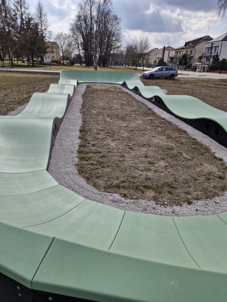 Zielony, falisty tor pumptrack otoczony jest żwirem. W tle stoją drzewa i budynki mieszkalne. Niebo zachmurzone z przebłyskami słońca.