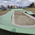 20260319_115559 Na zdjęciu widoczny jest zielony tor pumptrack na otwartym terenie. W tle widać domy, drzewa i niebieski samochód. Na niebie znajdują się białe chmury.