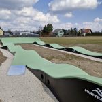 20260319_115609 Tor pumptrack o zielonej nawierzchni i czarnych bokach na żwirowej ścieżce. W tle widoczne są domy i drzewa pod niebieskim niebem z chmurami.