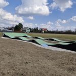 20260319_115620 Na obrazie widać terenową trasę rowerową o falistym kształcie w zielonym i czarnym kolorze. W tle znajdują się domy, drzewa i niebieskie niebo z białymi chmurami. Na pierwszym planie widoczna jest ziemia bez roślinności