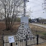 20260325_094712 Kamienny pomnik z metalowym krzyżem otoczony jest czarnym ogrodzeniem. W tle drzewa i niebo, po lewej strona jest tablica. Obok pomnika znajduje się tablica z informacjami.