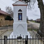 20260326_144421 Biała kapliczka z niebieską figurą na tle wiejskiego krajobrazu. Otoczona czarnym ogrodzeniem, obok rośnie duże drzewo. Na ziemi kamienna posadzka i kwiaty w donicy.