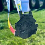 Medal w kształcie mapy z napisem II Bieg Konstantego, 25 kwietnia 2026. W tle zielona trawa i drzewa. Wstążka medalu jest biało-niebieska z żółtymi i czerwonymi akcentami.