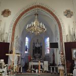 5sTqCSv3 Wnętrze kościoła z ołtarzem, białymi ścianami i złotym żyrandolem. Witraże i religijne obrazy na ścianach. Dekoracje kwiatowe w bieli i czerwieni.