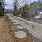 Droga w remoncie, pokryta piachami i zaprawą, otoczona drzewami. Po prawej stoi ozdobiona kapliczka. Tło to jasne, zachmurzone niebo.