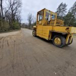 Żółty wózek widłowy stoi na nieutwardzonej drodze. W tle widać drzewa bez liści i szare niebo. Teren jest pusty i naturalny.