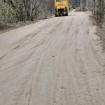 Na wąskiej, piaszczystej drodze stoi żółty walec. Wokół drzewa z młodymi listkami. W tle widać zalesiony teren.