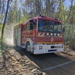 Czerwony wóz strażacki jedzie po leśnej drodze, wzbijając kurz. W tle zielone drzewa pod błękitnym niebem. Na pojeździe widoczny jest numer 517O21.