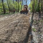 Droga gruntowa w lesie jest równana przez pomarańczowy spychacz. W tle drzewa i niebo, osoba stoi obok maszyny. Dominują brązy i zielenie.