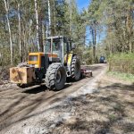 Traktor na leśnej drodze wśród drzew. Jasnoniebieskie niebo, ziemia przygotowywana do wyrównania. W tle bardziej zalesiony krajobraz.