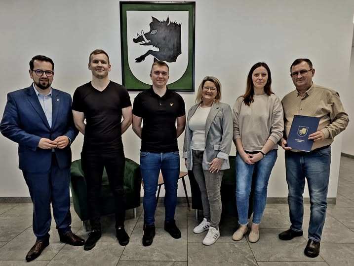 Sześć osób stoi przed białą ścianą z czarno-białym herbem dzika. Ubrania w odcieniach granatu, czerni i szarości. Jeden z mężczyzn trzyma niebieską teczkę.