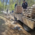 Ciężarówka z workami cementu na leśnej drodze, mężczyźni rozładowują cement. Tło to zielone drzewa i błękitne niebo. Ziemia jest mokra i błotnista.