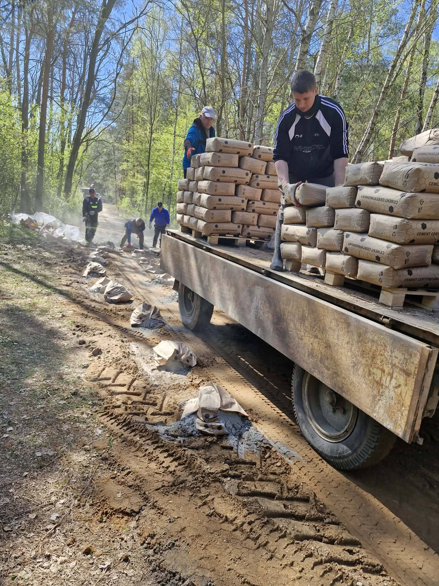 Ciężarówka z workami cementu na leśnej drodze, mężczyźni rozładowują cement. Tło to zielone drzewa i błękitne niebo. Ziemia jest mokra i błotnista.