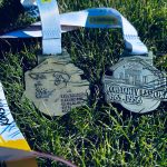 Dwa srebrne medale leżą na zielonej trawie. Mają wypukłe wzory i białe wstążki z kolorowymi napisami. Tło jest żywo zielone.