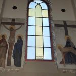 7SdS4V12 Na obrazie Jezus na krzyżu i scena zdjęcia z krzyża. Kolory dominujące to brązowy i niebieski. Tło to duże okno z witrażem.