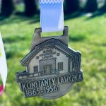 Metalowy medal z budynkiem, imieniem Konstanty Laszczka i datami 1865-1956. Srebrny kolor, w oddali zielone tło.