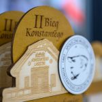 Trofeum z drewna z napisem II Bieg Konstantego i medalem z twarzą na tle. Kolory beżowy i biały, rozmyte tło.