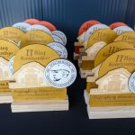Na zdjęciu widać drewniane trofea z napisem II Bieg Konstantego na czarnym stole. Trofea mają okrągły emblemat z rysunkiem budynku i portretem. W tle widoczne są podobne nagrody w kolorach żółtym, pom