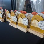 Na stole stoją drewniane trofea w różnych kolorach z napisami i białymi medalami. Tło jest ciemne z zasłoną. Trofea mają napisy o II Biegu Konstantego.