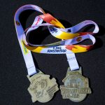 Dwa medale z białymi wstążkami na czarnym tle. Na jednym medal z twarzą, na drugim budynek i napis Konstanty Łazicki. Kolory wstążek to biały, czerwony i żółty.