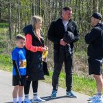 Na zdjęciu jest grupa czterech osób na ścieżce, na tle zieleni i drzew. Mężczyźni są ubrani na ciemno, chłopiec ma niebieską koszulkę. Obok nich jest kobieta w czarnej kurtce, rozmawiają.