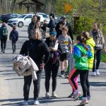 Grupa osób spaceruje po asfaltowej drodze, otoczonej zielenią. Ludzie ubrani są głównie w sportowe ubrania i kurtki. W tle widać zaparkowane samochody i drzewa.