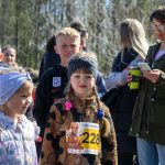 Grupa dzieci stoi na zewnątrz, niektóre mają numery startowe. Tło to drzewa, a jedna osoba trzyma telefon. Kolory są jasne, wiosenne.