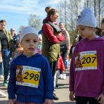 Na zdjęciu widoczne dzieci w numerach startowych uczestniczą w biegu. Mają na sobie kolorowe bluzy i czapki. Tło stanowi tłum osób na tle drzew.