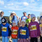 Dzieci w kolorowych ubraniach z numerami startowymi stoją na zewnątrz. Tło to niebo z chmurami i dorośli w tle. Jest słoneczna pogoda.