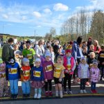 Grupa dzieci w kolorowych ubraniach czeka na start biegu. Na tle widoczne są drzewa i niebo z chmurami. Obok stoi zegar elektroniczny.