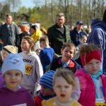 Grupa dzieci w kolorowych ubraniach stoi na zewnątrz. W tle widać drzewo i rozmyte postacie dorosłych. Dzieci mają numerki na koszulkach, sugerujące udział w biegu.