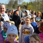 Grupa ludzi na zewnątrz przy słonecznej pogodzie. Kobieta z kijkiem oraz dzieci w kolorowych ubraniach. Tło: drzewa, samochody.