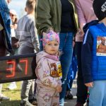 Na zdjęciu jest małe dziecko w różowej czapce i bluzie z postaciami z bajek. W tle grupa ludzi na zewnątrz. Obok widoczny jest licznik z numerem 37.