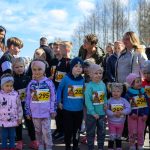 Grupa dzieci w kolorowych ubraniach stoi na zewnątrz z numerami startowymi. Tło stanowią drzewa i niebieskie niebo. W tle dorośli obserwują wydarzenie.