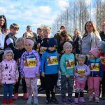 Grupa dzieci i dorosłych stoi na zewnątrz w słoneczny dzień, dzieci mają numery startowe. Tło to błękitne niebo z chmurami. W tle widoczny jest rząd drzew.