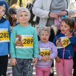 Grupa dzieci w kolorowych ubraniach z numerami startowymi stoi na zewnątrz. Tło jest rozmyte, co podkreśla głównych bohaterów. Dominują odcienie niebieskiego, różowego i turkusowego.