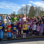 Na zdjęciu widać grupę dzieci w kolorowych strojach sportowych na linii startu. Dzieci trzymają numery startowe na jasno oświetlonej drodze. W tle jest zielony las i niebieskie niebo z chmurami.