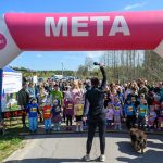 Pod dużym różowo-niebieskim łukiem startowym META zebrana grupa dzieci i dorosłych. Na pierwszym planie osoba z mikrofonem. Pogodny dzień z niebieskim niebem w tle.