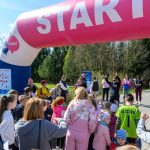 Grupa ludzi gromadzi się pod dużym różowym łukiem z napisem START. W tle widać drzewa i niebo. Uczestnicy są w różnorodnych kolorowych strojach, gotowi do biegu.