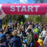 Na zdjęciu widoczny jest tłum ludzi na starcie pod różowo-białą bramką. W tle drzewa i niebo z chmurami. Uczestnicy ubrani są w kolorowe ubrania, a atmosfera jest radosna.