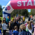 Grupa ludzi, głównie dzieci, stoi na starcie biegu pod różowo-niebieskim łukiem. Tło to zielone drzewa i błękitne niebo. Atmosfera jest radosna i pełna oczekiwania.