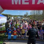 Dzieci zgromadzone na starcie wyścigu pod różowo-białym balonem z napisem META. Słoneczny dzień, zielone drzewa w tle. Elektroniczny zegar wskazuje czas.