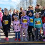 Grupa dzieci stoi na starcie maratonu, w jasnych kolorowych ubraniach i numerach startowych. Tło to drzewa i niebo. Obraz emanuje energią i radością.