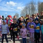 Dzieci startują w biegu, ubrane kolorowo, z numerami na koszulkach. Tło stanowi niebo i drzewa. Słoneczna pogoda.