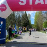 Start dziecięcego biegu pod różowo-białą bramką z napisem. W tle zielone drzewa i niebieskie niebo. Kibice stoją po bokach.