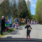 Dziewczynka w czarnym stroju biegnie do mety na drodze. W tle widoczni kibice, drzewa i niebieskie niebo. Atmosfera sportowa i radosna.