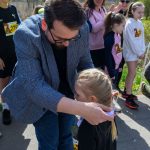 Mężczyzna w niebieskiej marynarce wręcza medal dziewczynce w czarnym stroju. W tle inne dzieci i dorośli, przyroda. Słoneczny dzień.