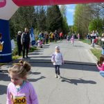 Grupa dzieci biegnie obok dmuchanej bramy na asfaltowej drodze. Po bokach są widzowie, a zegar pokazuje 10:25. Tło stanowią drzewa i niebo.